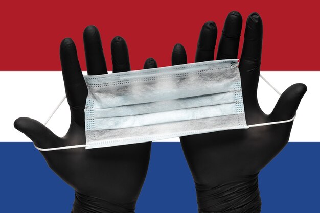 Doctor holds face mask in hands in black medical gloves on background colors flag of netherlands con...