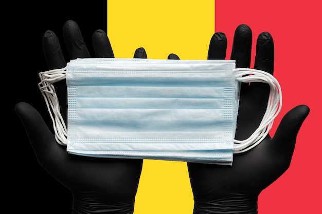 Doctor holds medical face mask respiratory bandage in hands in black gloves on background flag of be...