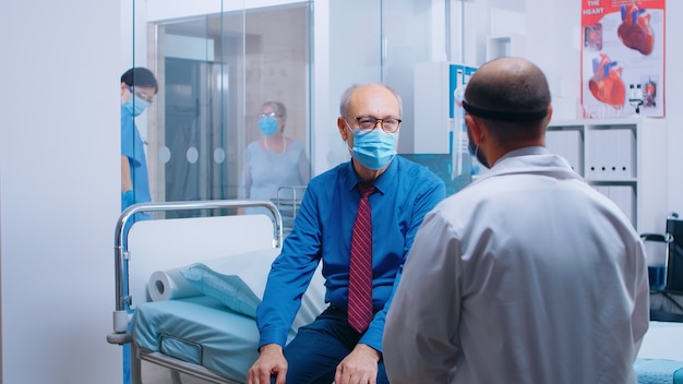Консультация врача в больнице во время глобальной пандемии COVID-19. Старый пенсионер старший мужчина в маске и медицинский работник в защитном снаряжении для консультации. Современная частная клиника