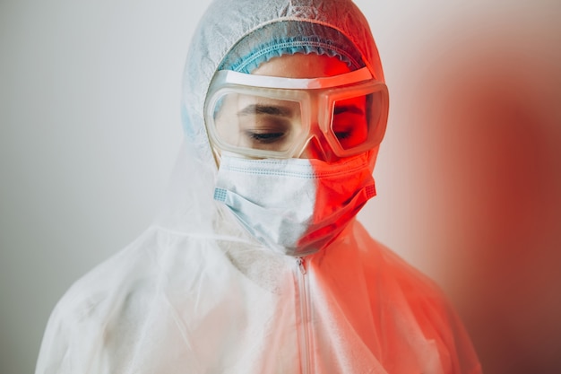 Doctor in beschermende uniform, bril, handschoenen op een blauwe achtergrond in neonlicht. close-up portret van een arts in rood neon. moe man vecht tegen een coronavirus. COVID 19