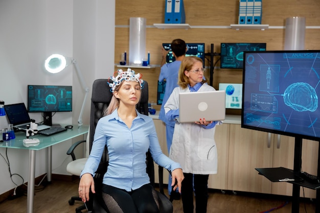 Doctor introducing some data into the laptop during a girl's brain scan procedure. Brain waves scanning helmet