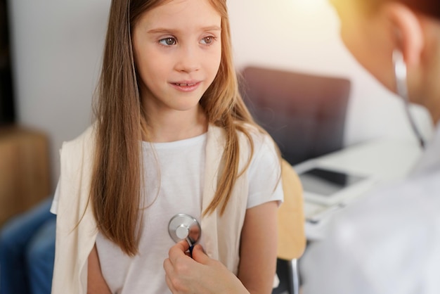 Doctor and kid patient are in the clinic. Physician in white coat examining a serious young girl with a stethoscope, close up. Medicine, therapy concept