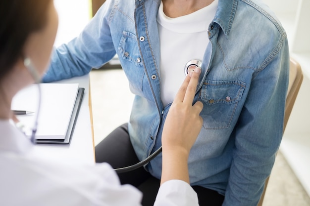 Doctor listening to cheerful young patients chest with stethoscope in his office at the hospital