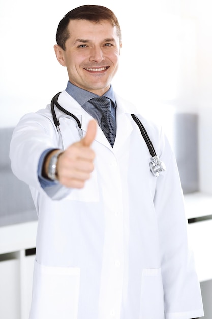 医者の男はカメラに向かって元気に笑って、親指を立てます。クリニックでの完璧な医療サービス。医学とヘルスケアの幸せな未来。