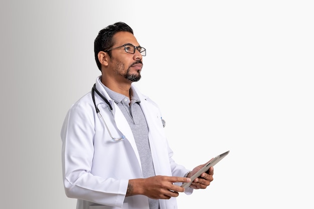 doctor, man holding tablet looks up distantly, wearing white uniform and stethoscope, white backgrou