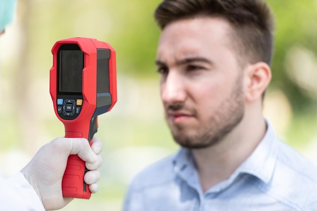 Doctor Man Use Infrared Forehead Thermometer Gun to Check Body Temperature for Virus Covid-19 Symptoms With the Isolation Gown or Protective Suits and Surgical Face Masks Outdoors in Park