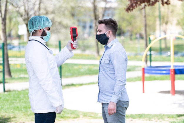 Photo doctor man use infrared forehead thermometer gun to check body temperature for virus covid-19 symptoms with the isolation gown or protective suits and surgical face masks outdoors in park