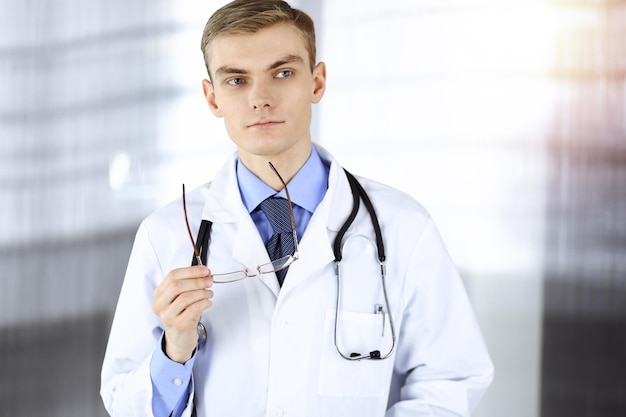 Doctor man, with glasses and stethoscope, is standing at his sunny hospital office. Perfect medical service in clinic. Happy future in medicine and healthcare.