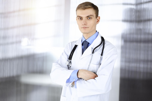 Doctor man, with a stethoscope, stands at his sunny hospital office. Perfect medical service in clinic. Happy future in medicine and healthcare.