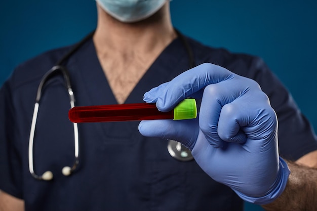 마스크를 쓴 의사, 파란색 일회용 장갑, 의료 스크럽, phonendoscope. 그의 손에 녹색 모자와 함께 빨간색 실험실 테스트 튜브를 들고. 파란색 배경입니다. 코로나바이러스 연구. 코로나 바이러스 감염증 -19 : 코로나 19. 클로즈업, 복사 공간