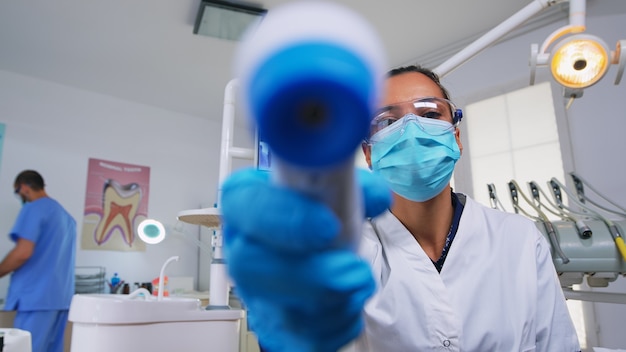 Foto medico che misura la temperatura della donna prima dell'esame odontoiatrico, paziente pov. dentista e infermiere che lavorano in un moderno ufficio ortodontico, scrivendo ed esaminando la persona che indossa una maschera di protezione