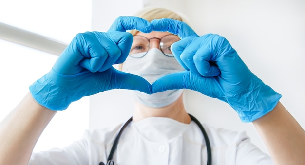 Doctor in a medical coat, mask and stethoscope showing heart in hands