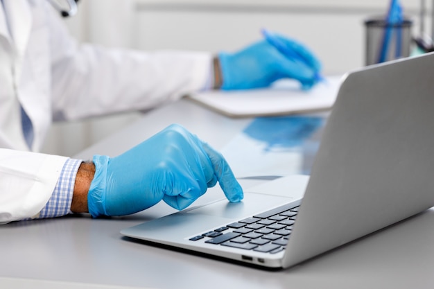 Doctor in medical gown and gloves typing on laptop and making notes at working table close up