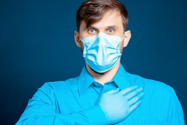 A doctor in a medical mask and gloves, in a blue uniform, put his hand to his chest.