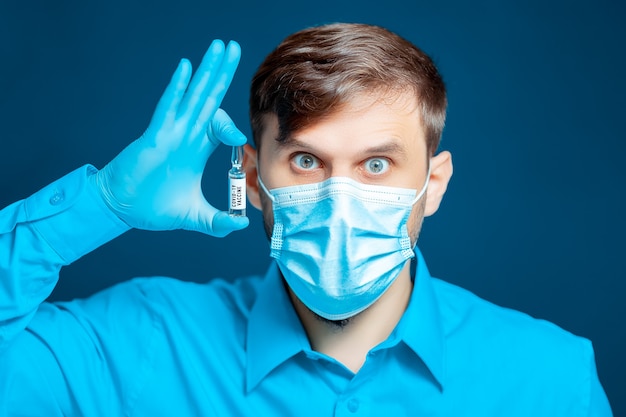 Doctor in a medical mask and gloves, dressed in a blue uniform holds the coronavirus vaccine near his eye