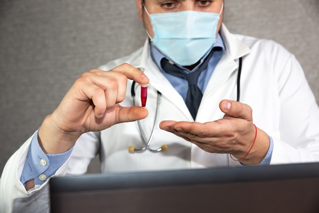 노트북 근처 의료용 마스크를 쓴 의사. 코로나바이러스 전염병 동안 원격 치료. 일반 개업의는 피가 든 유리병을 들고 있습니다. 분석 중인 바이러스. 온라인 컨설팅.