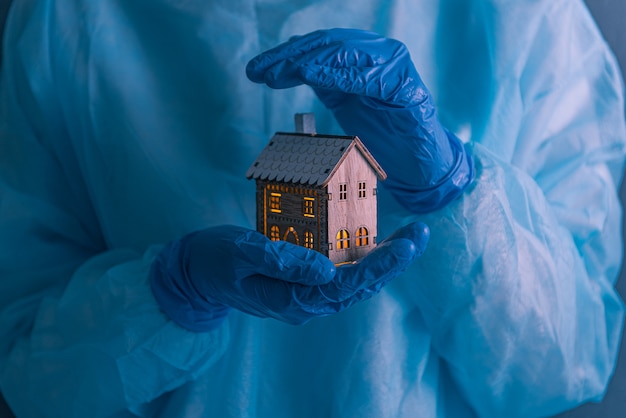 Доктор, медицинский работник с медицинскими перчатками на руках, пальто и маской на лице, держит в руках маленький домик со светящимися окнами.