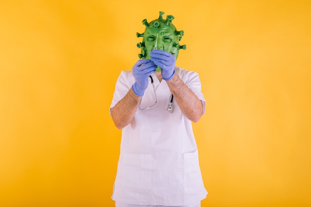 Doctor - nurse disguised as coronavirus wearing latex mask - covid-19 virus with a syringe with water in her hands on yellow background. coronavirus concept