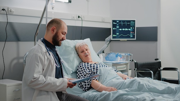 Doctor and nurse doing consultation for sick patient in bed. Medical team checking healthcare treatment of retired woman in hospital ward with nasal oxygen tube and IV drip bag.