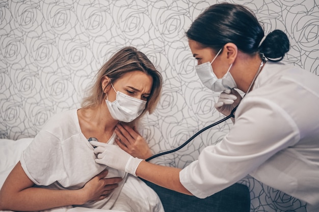 Photo doctor nurse in protective face mask listening to breath with a stethoscope suspecting coronavirus (covid-19). first symptoms concept. woman sick of flu viral infection in home isolation quarantine