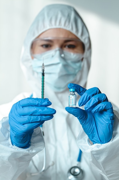 Doctor or nurse in a protective suit holds a syringe and a covid 19 vaccine