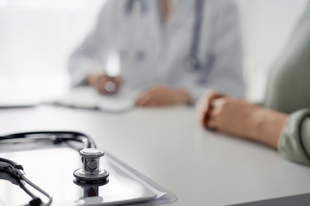 Doctor and patient are sitting and discussing something at the desk in the clinic office. The focus is on the stethoscope lying on the table, close up. Perfect medical service and medicine concept.