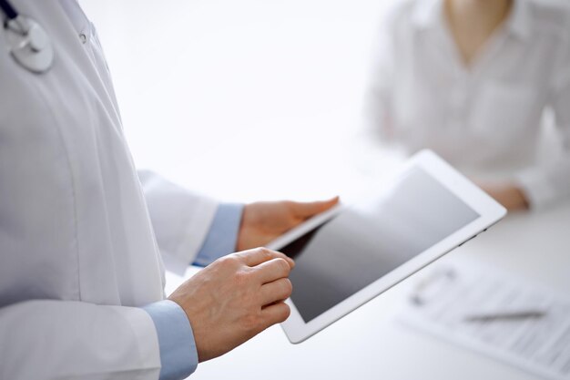 Doctor and patient in clinic. The focus is on female physician's hands using tablet computer at the background of unknown woman. Medicine concept.