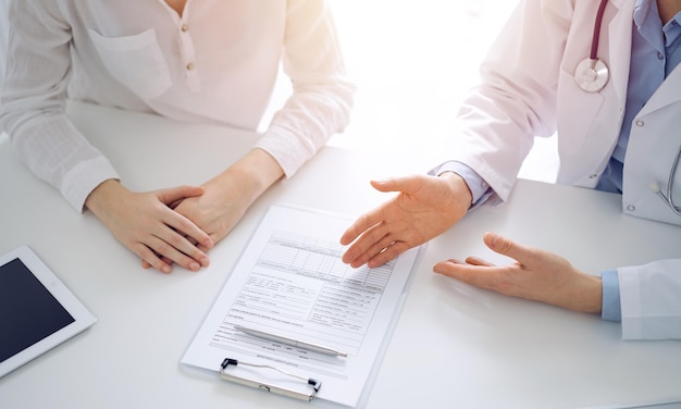 Doctor and patient discussing current health questions while sitting near of each other and using clipboard at the table in clinic, just hands closeup. Medicine concept