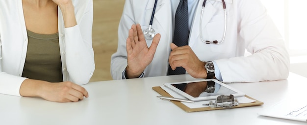 Doctor and patient discussing medical exam results while sitting at the desk in sunny clinic closeup Male physician using tablet computer for filling up medication history record