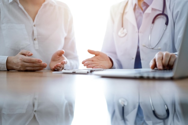 Doctor and patient discussing something while using laptop computer and sitting near each other at the wooden desk in clinic. Medicine concept.