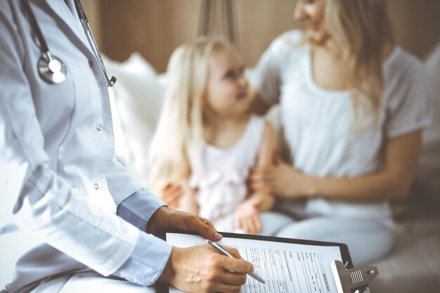 Doctor and patient. Pediatrician using clipboard while examining little girl with her mother at home. Happy cute caucasian child at medical exam. Medicine concept.