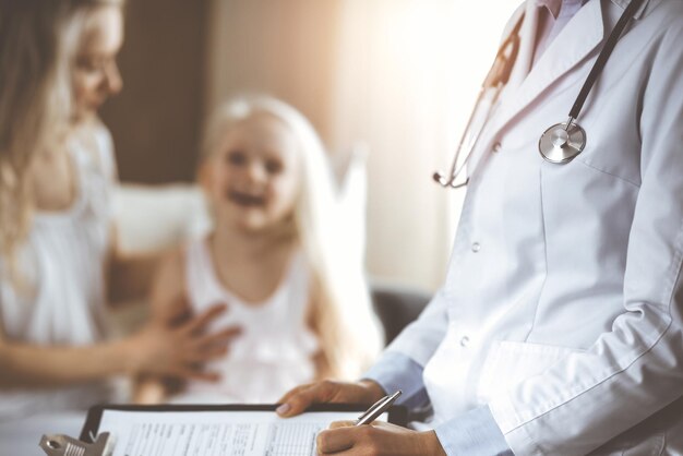 Doctor and patient. Pediatrician using clipboard while examining little girl with her mother at home. Happy cute caucasian child at medical exam. Medicine concept.