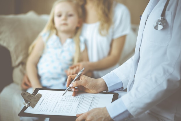 Doctor and patient. Pediatrician using clipboard while examining little girl with her mother at home. Sick and unhappy child at medical exam. Medicine concept.