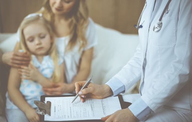 Doctor and patient. Pediatrician using clipboard while examining little girl with her mother at home. Sick and unhappy child at medical exam. Medicine concept