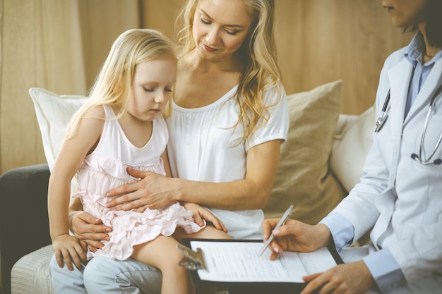 Doctor and patient. Pediatrician using clipboard while examining little girl with her mother at home. Sick and unhappy child at medical exam. Medicine concept.