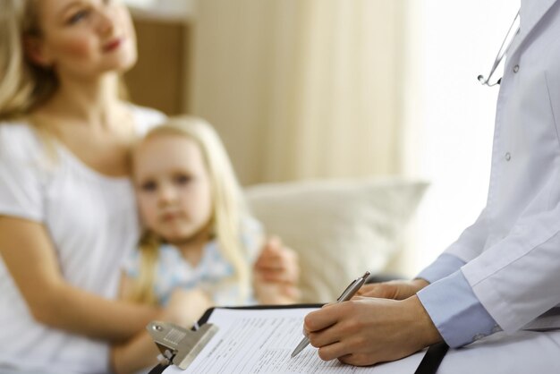 Doctor and patient. Pediatrician using clipboard while examining little girl with her mother at home. Sick and unhappy child at medical exam. Medicine concept.