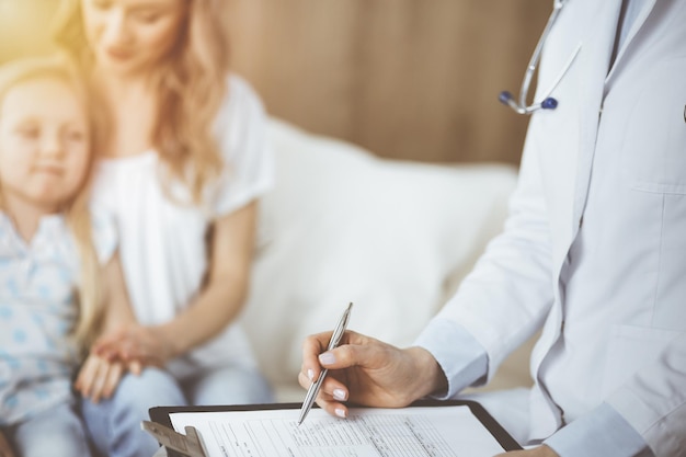 Doctor and patient. Pediatrician using clipboard while examining little girl with her mother at home. Sick and unhappy child at medical exam.