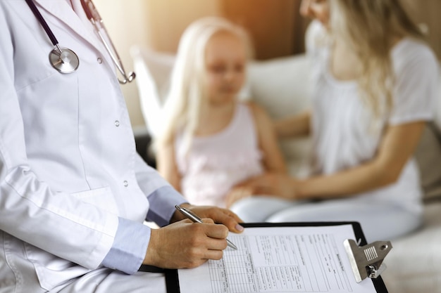 Doctor and patient. Pediatrician using clipboard while examining little girl with her mother at home. Sick and unhappy child at medical exam.