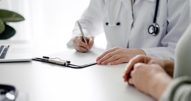 Doctor and patient sitting at the desk in clinic office. The focus is on female physician's hands filling up the medication history record form, close up. Medicine concept.