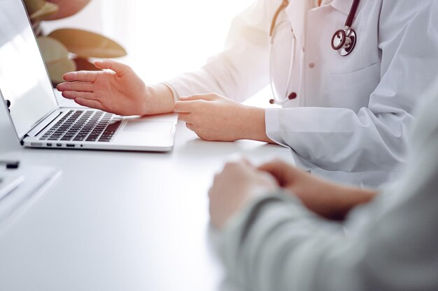 Doctor and patient sitting at the desk in clinic office. The focus is on female physician's hands pointing to laptop computer monitor, close up. Medicine concept