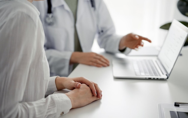 Doctor and patient sitting at the desk in clinic office. The focus is on female physician's hands pointing to laptop computer monitor, close up. Perfect medical service and medicine concept