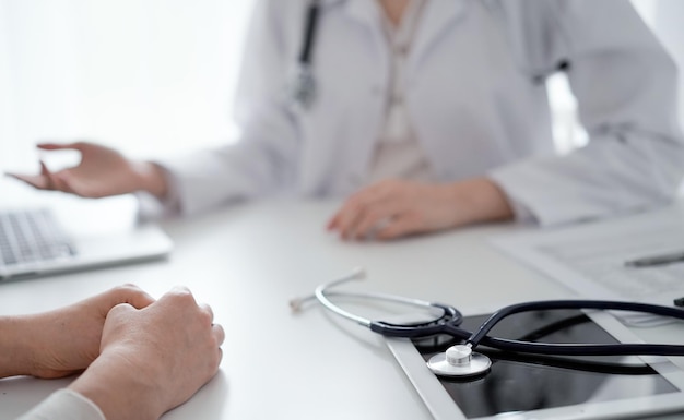 Doctor and patient sitting and discussing something at the desk in clinic. The focus is on the stethoscope lying on the table, close up. Medicine and healthcare concept