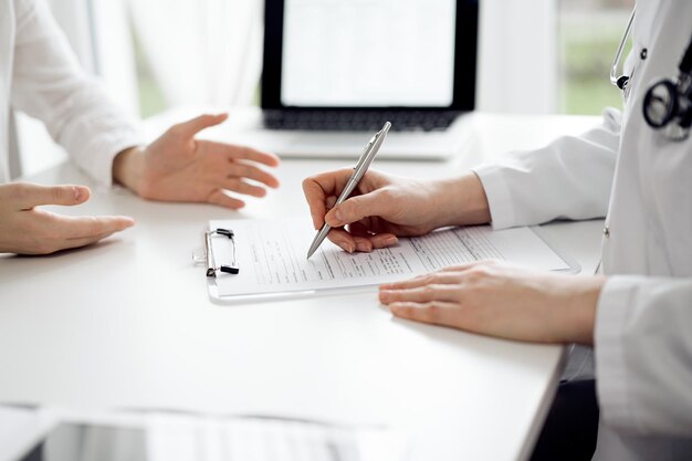 Doctor and patient sitting and discussing something near each other at the white desk in clinic. Female physician is listening filling up a records form. Medicine concept.