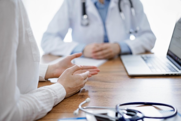 의사와 환자는 랩톱 컴퓨터를 사용하는 동안 나무 테이블에 앉아서 무언가를 논의합니다. 초점은 환자의 손에 있습니다. 의학 개념