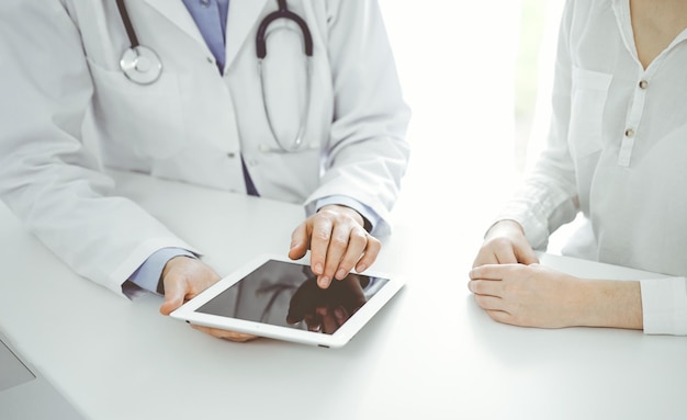 Doctor and patient sitting near each other at the desk in clinic. The focus is on female physician's hands pointing into tablet computer touchpad, close up. Medicine concept.