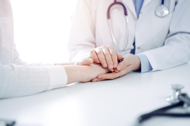 Doctor and patient sitting near each other at the table in clinic office. The focus is on female physician's hands reassuring woman, only hands, close up. Medicine concept