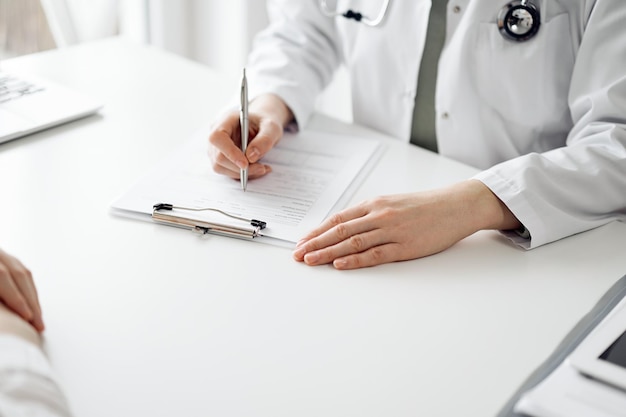 Doctor and patient sitting near each other at the white desk in clinic. Female physician is listening filling up a records form. Medicine concept