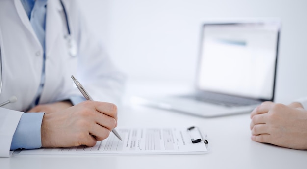 Doctor and patient sitting opposite each other at the desk in clinic. The focus is on female physician's hands filling up the medication history record form or checklist, close up. Medicine concept