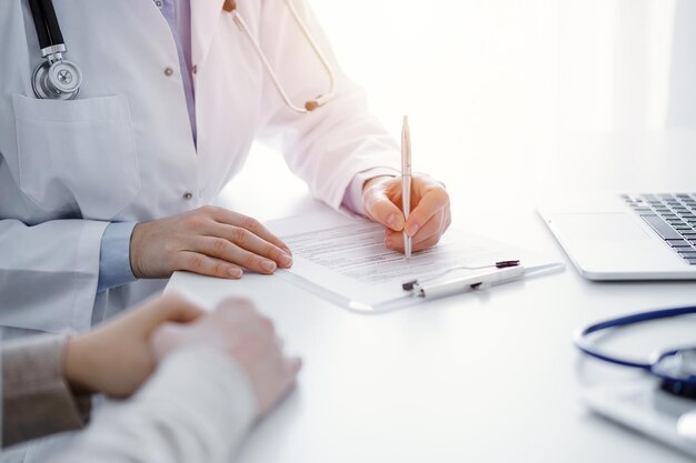 医師と患者が診療所のテーブルに座っています。薬歴記録フォームまたはチェックリストに記入する女性医師の手の接写に焦点が当てられています。医学の概念。