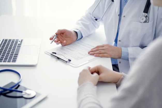 医師と患者が診療所のテーブルに座っています。薬歴記録フォームまたはチェックリストに記入する女性医師の手の接写に焦点が当てられています。医学の概念。